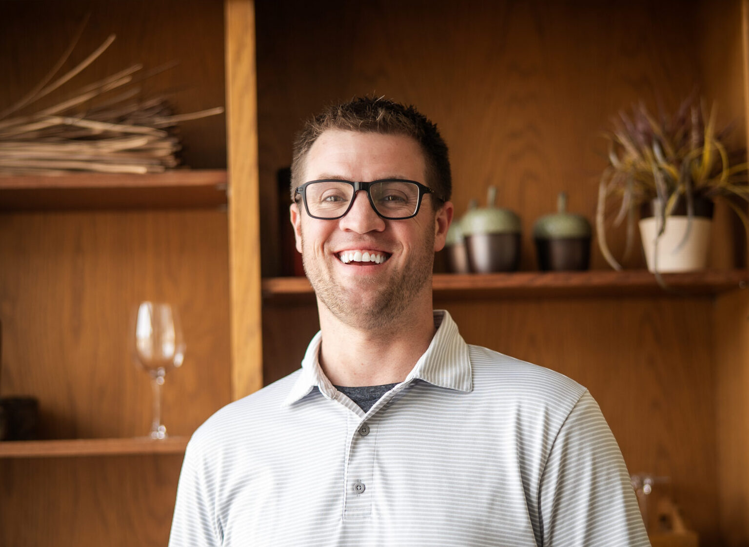 A fair skinned man in glasses and a light colored polo shirt smiles in front of a backdrop that includes a wooden shelf with various items on it.