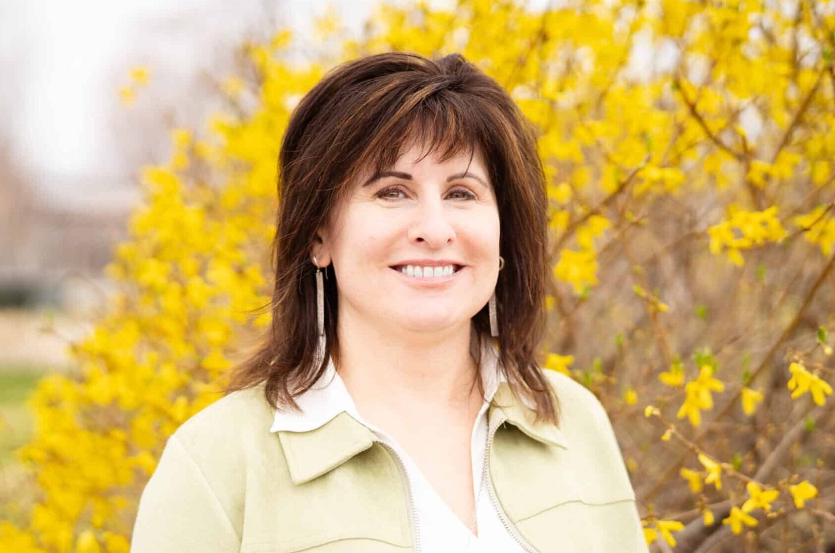 A smiling woman with brown hair and a light green jacket in front of a large bush with yellow blooms.