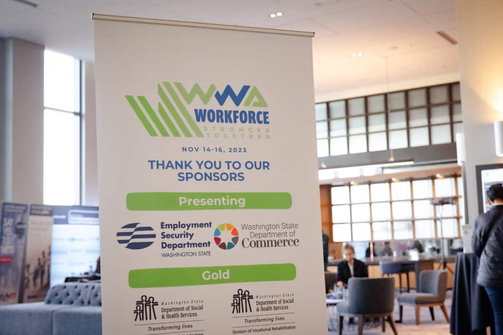 A standing vertical sign that says Thank You To Our Sponsors is in the front of a large and well lit conference room.