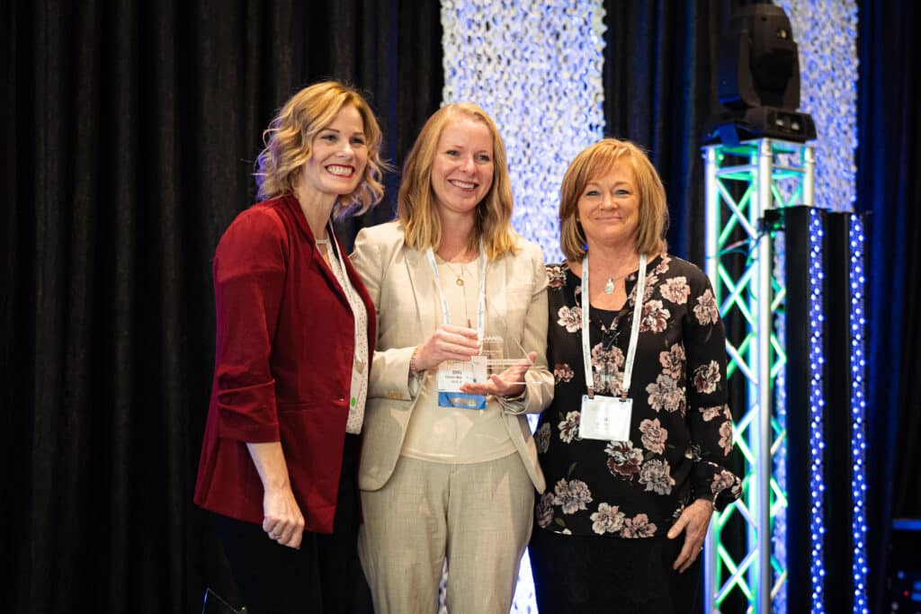 Amy Martinez in a red blazer and Lisa Romine in a black blouse with pink flowers, present a clear award to Chelsea Mason-Placek, who is smiling in between them and wearing an off white suit.