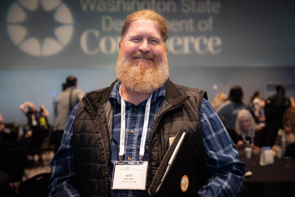 A red-haired, full-bearded Nate Mack smiles in front of a projected logo for Washington State Dept of Commerce.