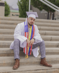 A male graduate in a white cap and gown sits on concrete steps.
