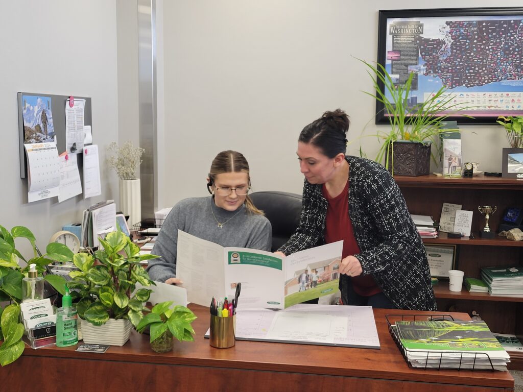 Two women look at a trifold document together behind a wooden desk with a few green plants on it.