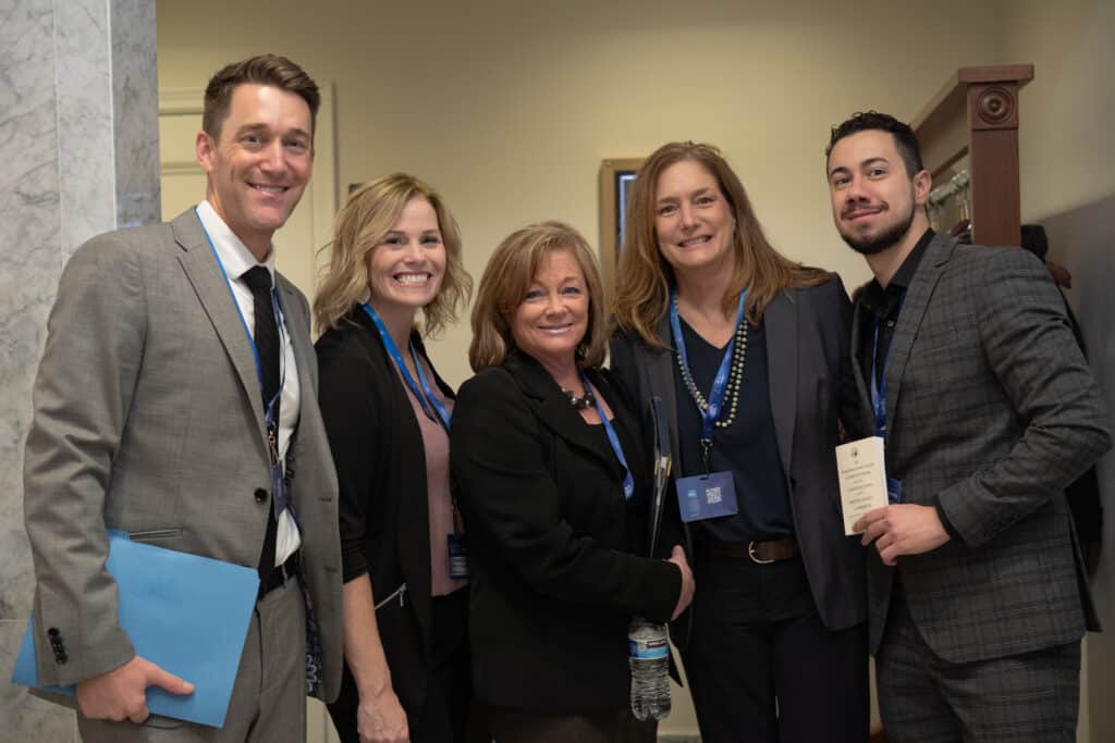 Five people in business wear stand in front of a Hearing Room at the Washington State Capitol. 