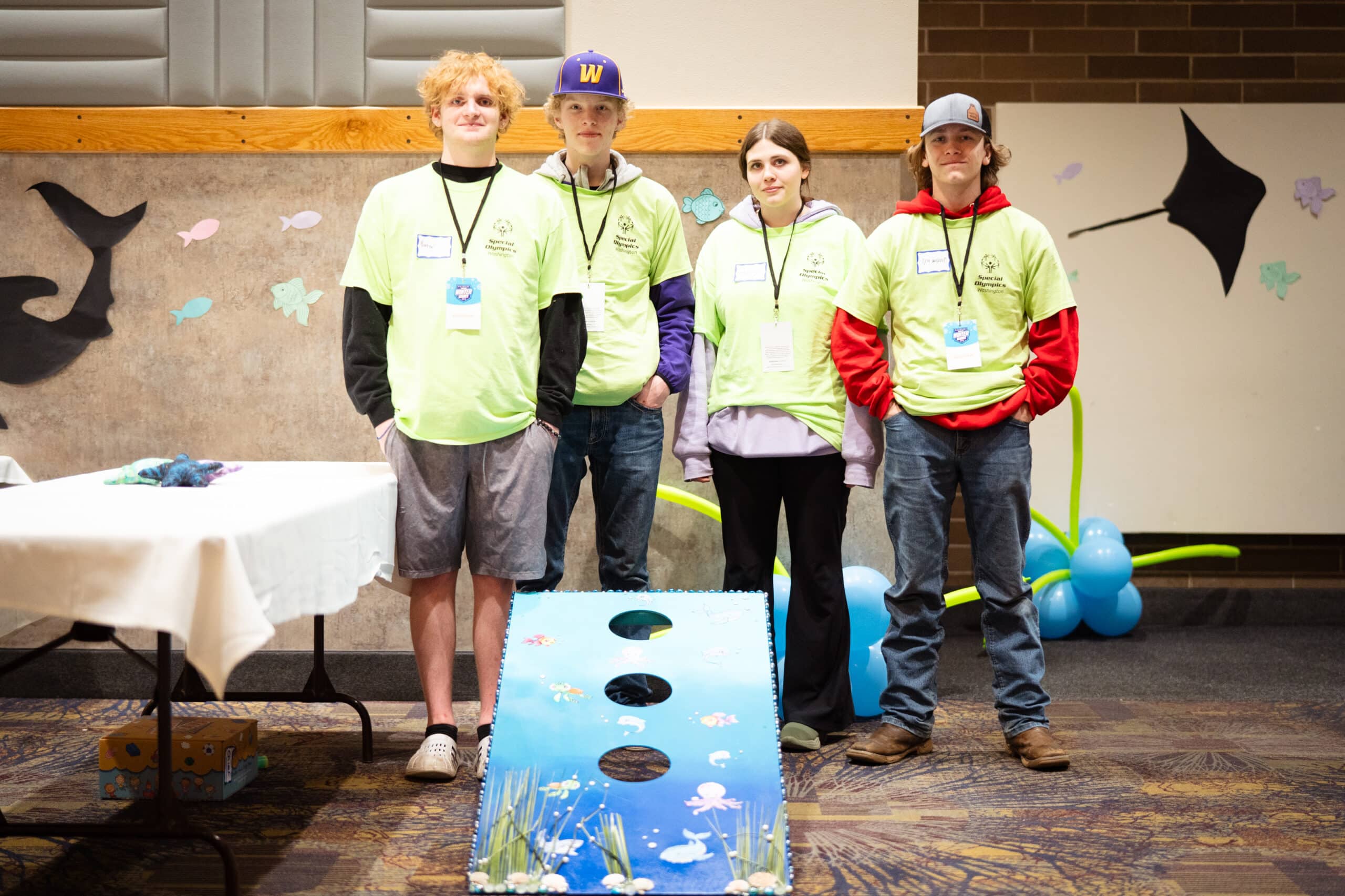Four students in yellow volunteer t-shirts stand behind their blue corn hole game that they made.