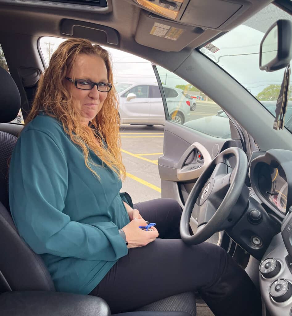 A woman in a blue shirt sits behind the steering wheel of a car that was just given to her.