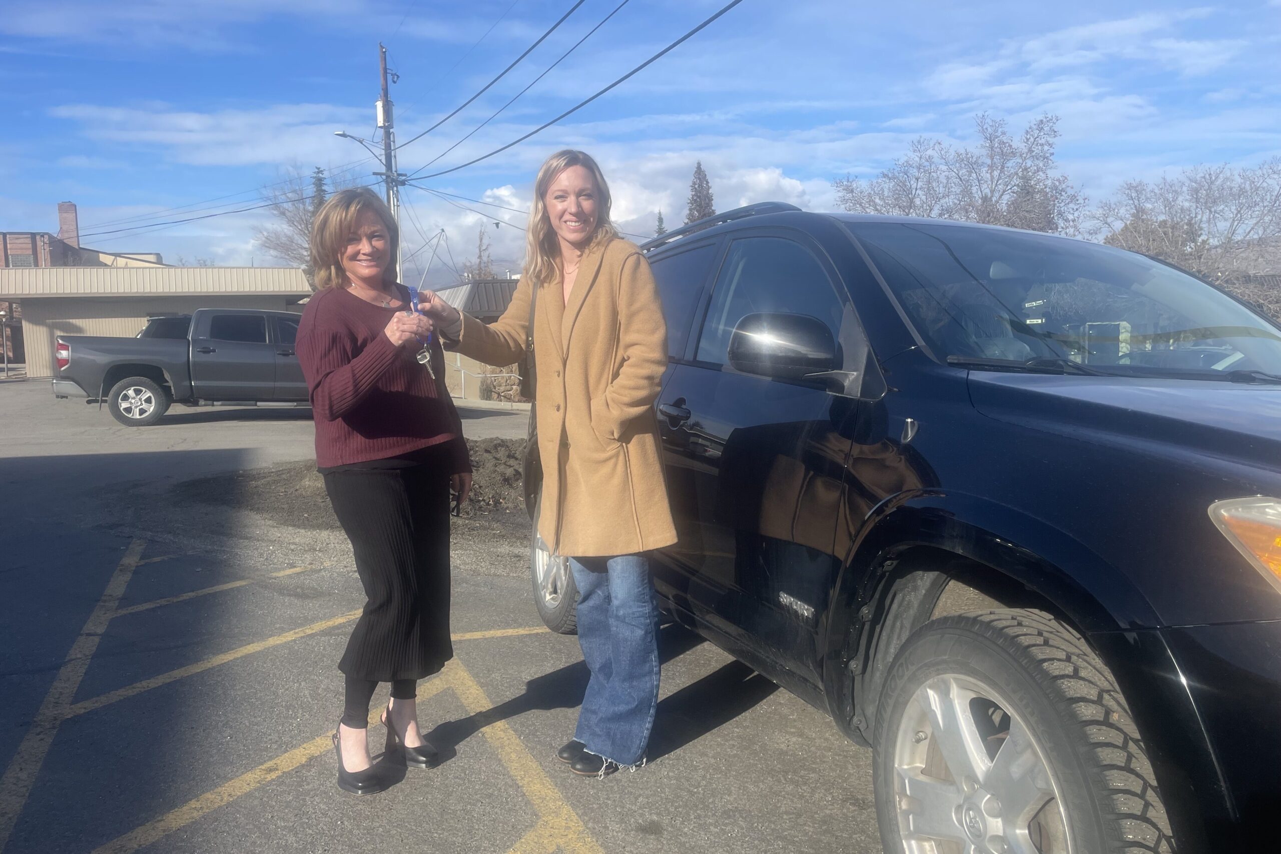 Two women stand in front of a black car as one gives the keys to the other.