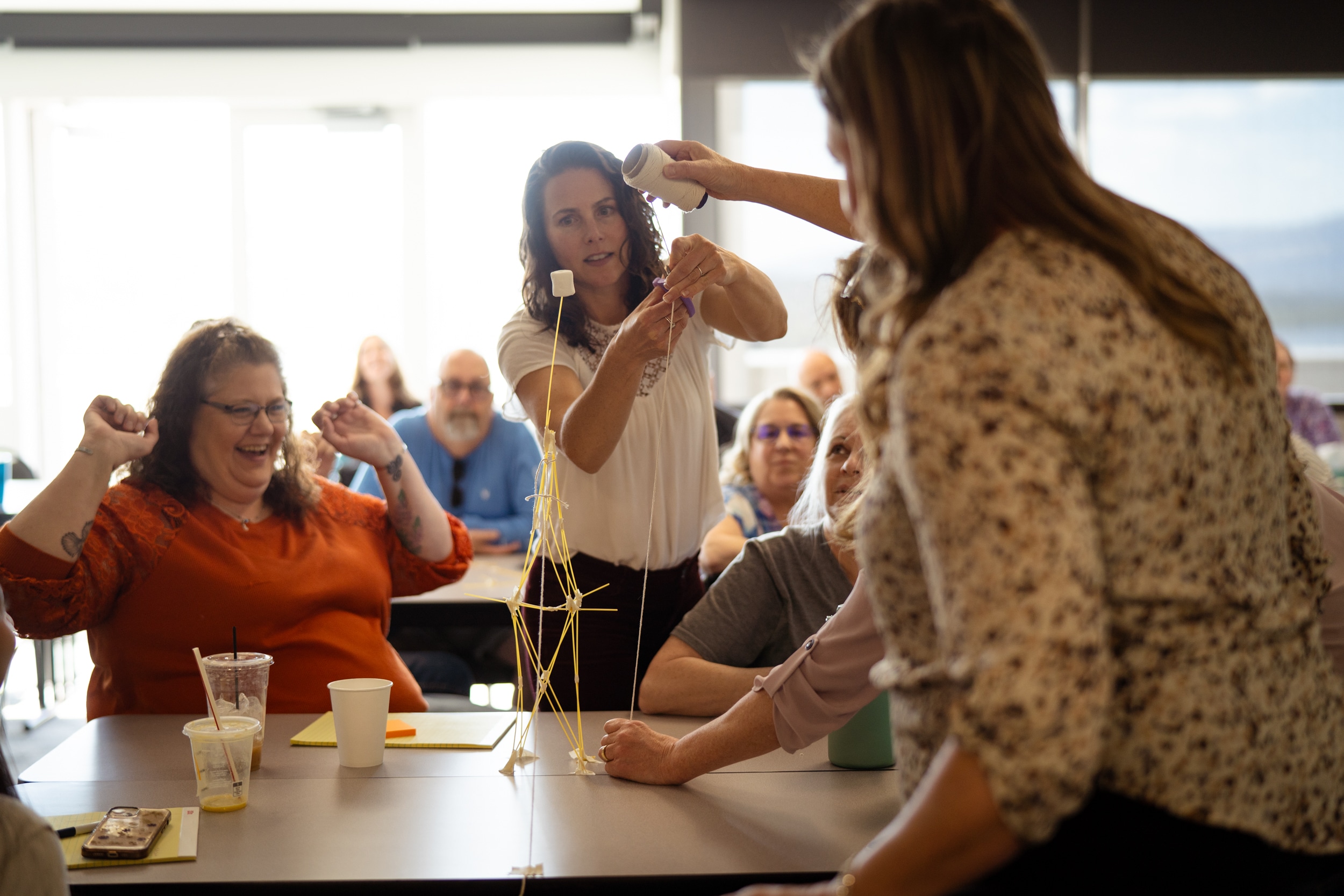A carefully constructed tower made of spaghetti with a marshmallow on top is measured by a woman in a white shirt while other people look on.