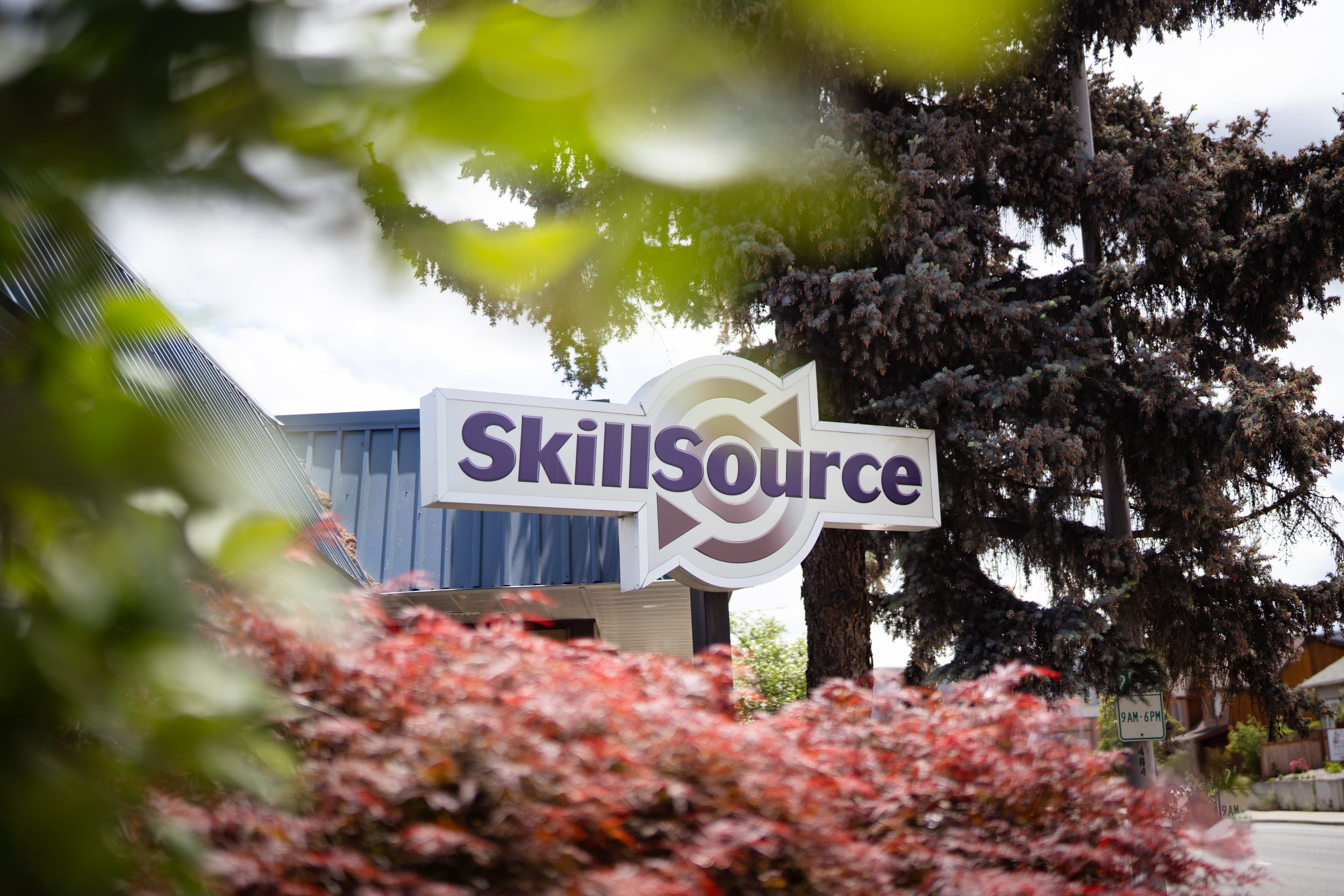 A freestanding sign on a pole that reads SkillSource is set back against a blue-roofed building and framed by green and burgundy colored foliage.