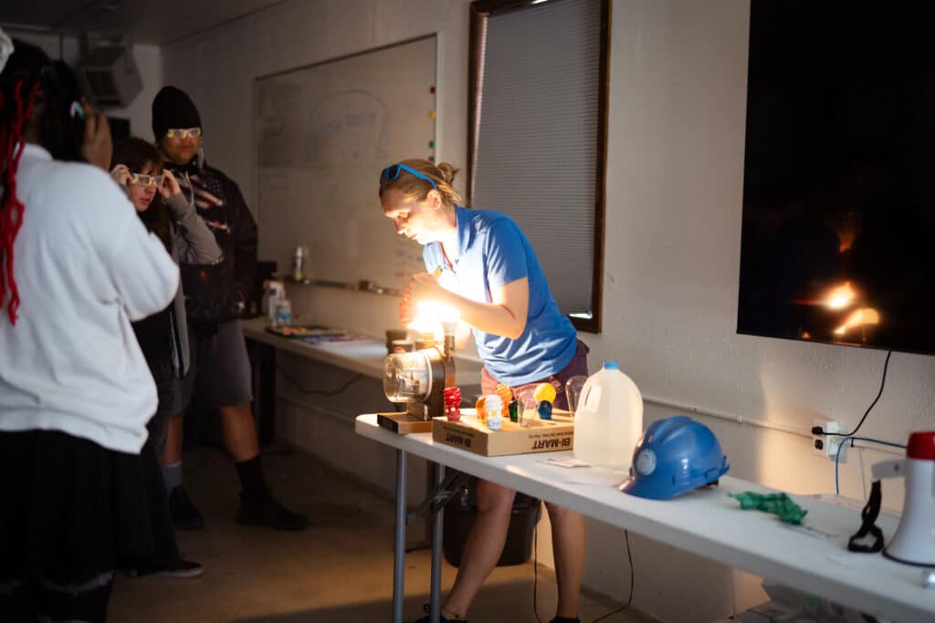 A female science teacher in a blue shirt holds her hands around a glowing light bulb in front of an audience of teen students.