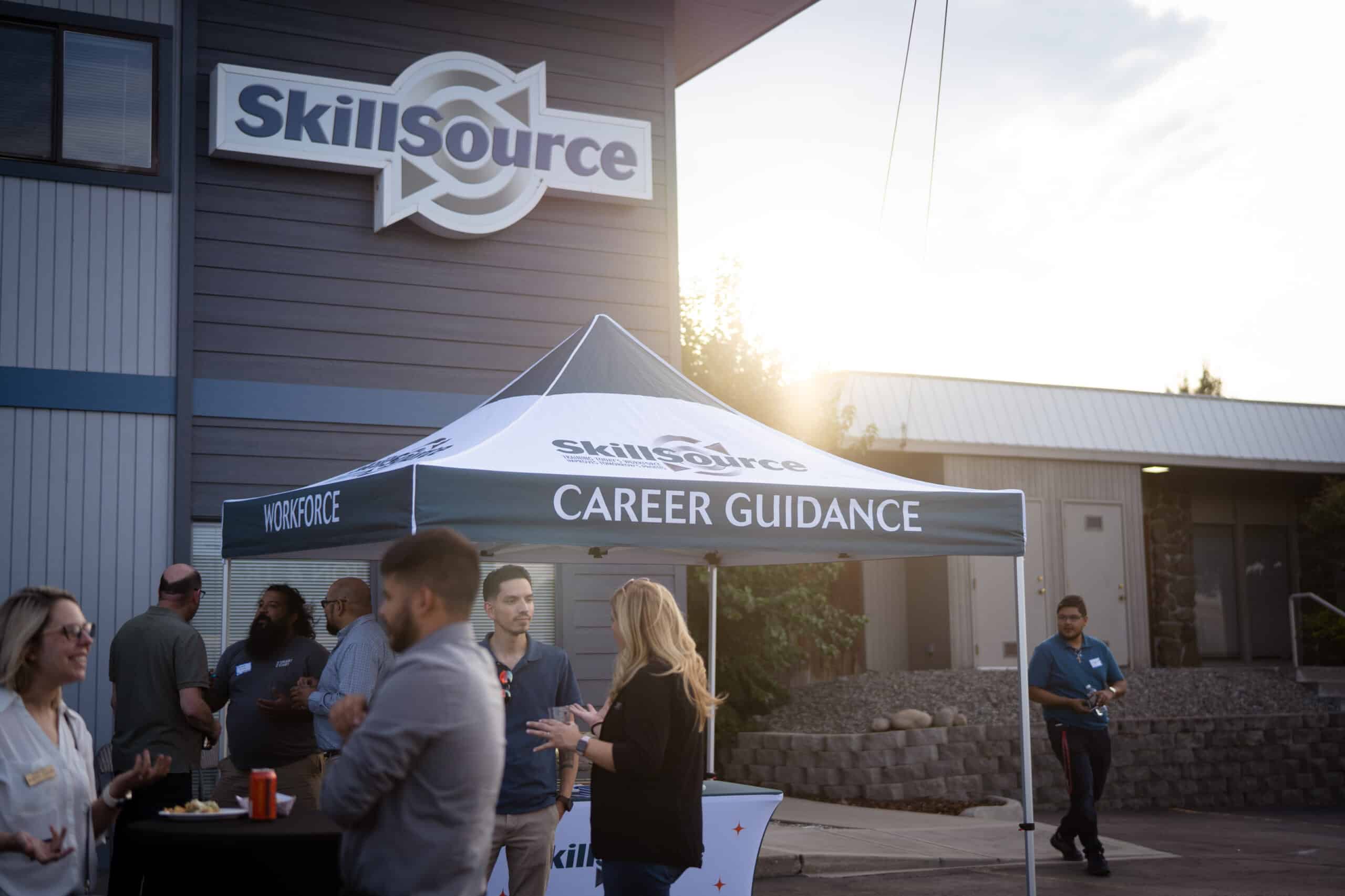 The sun sets behind a blue-gray building with a large sign on the exterior that says SkillSource and a pop-up tent out front with the words Career Guidance printed on it and smiling people milling around the tent and chatting.