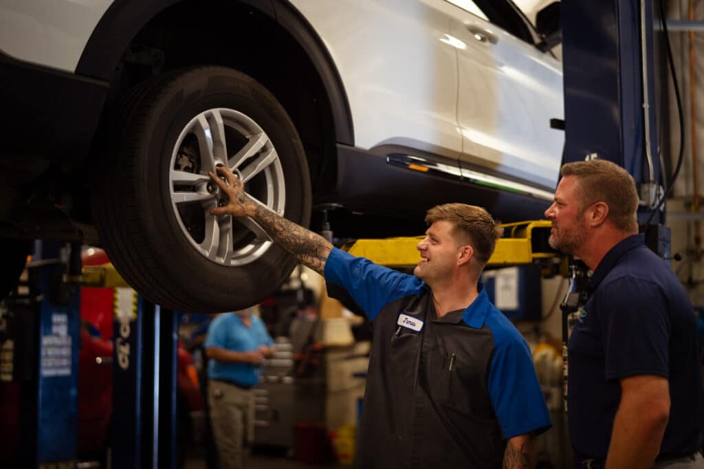 Two men with short hair and wearing dark blue work shirts inspect a tire on a vehicle that's up on a lift.