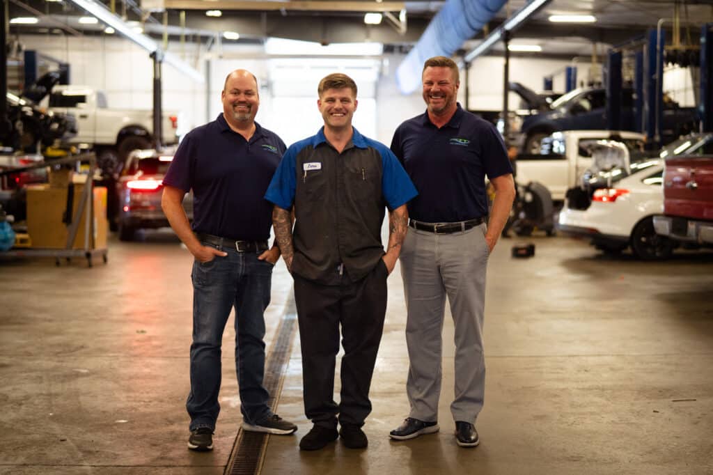 Three smiling men in dark blue work shirts stand side by side in the Ford automotive service area.