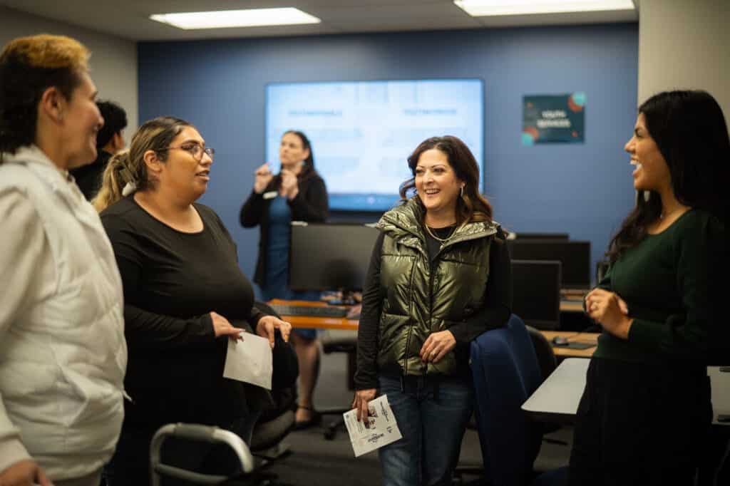A group of dark haired women talk and laugh while standing in a room where a presentation is being given.