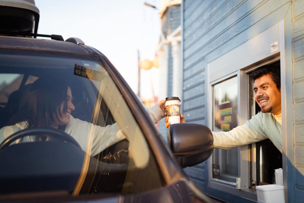 A latino man with dark hair and a light shirt smiles as he leans out of a drive through window and hands a cup of coffee to a smiling woman in her car.