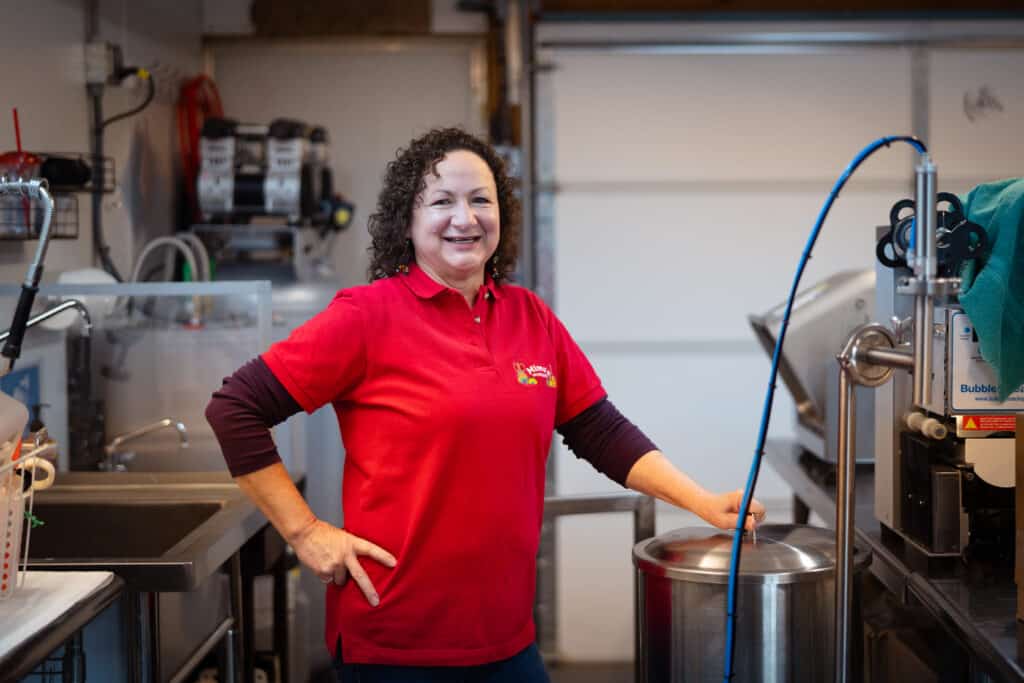 A smiling woman with dark curly hair wears a red shirt that bears the logo Mima's Famous Salsa and she stands next to an industrial filling machine.