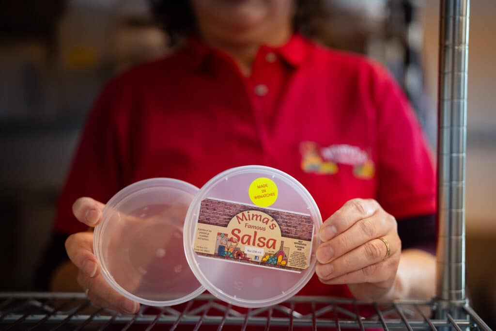 A woman in a red shirt holds in front of her an empty salsa container that bears the logo for Mima's Famous Salsa and includes a sticker that says Made in Wenatchee.