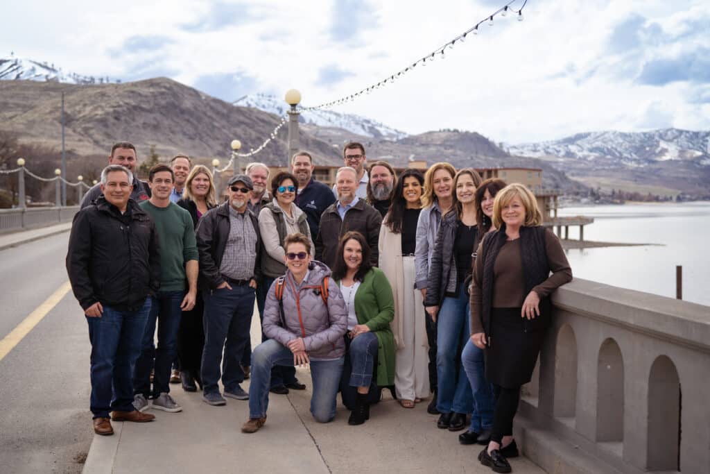 A diverse group of industry professionals gather for a group photo on a concrete bridge beneath a string of lights and against a backdrop of the lake and snowy hills.