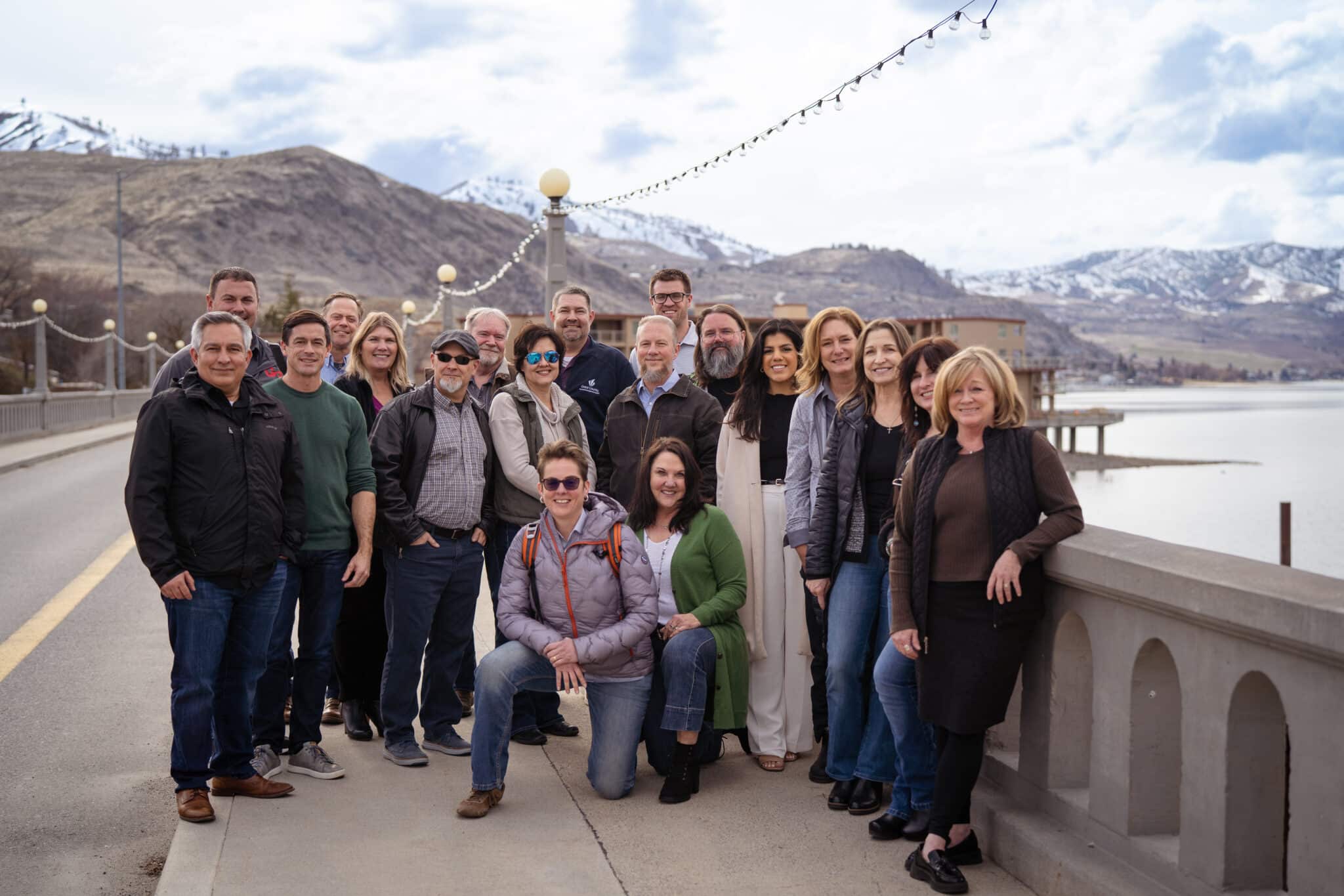 A diverse group of industry professionals gather for a group photo on a concrete bridge beneath a string of lights and against a backdrop of the lake and snowy hills.