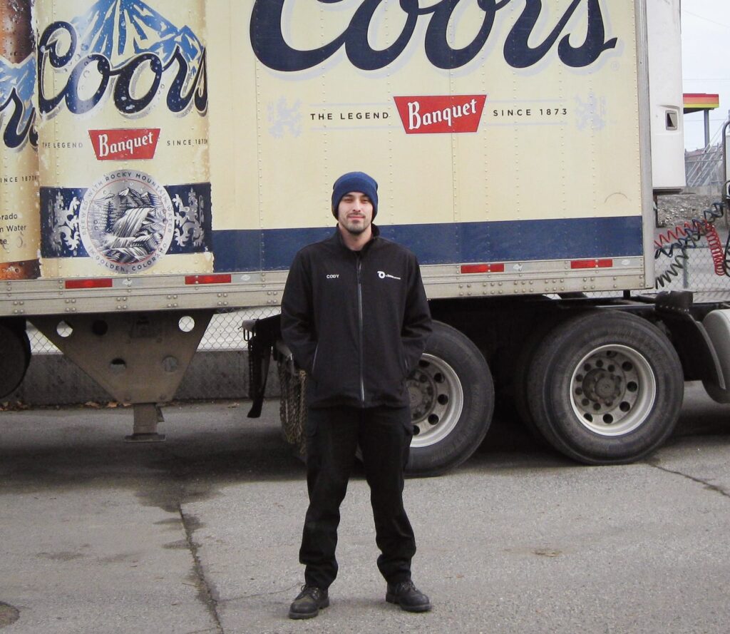 A man in black work clothes and a blue beanie cap stands with his hands in his pockets in front of a truck with the Coors logo on the side.