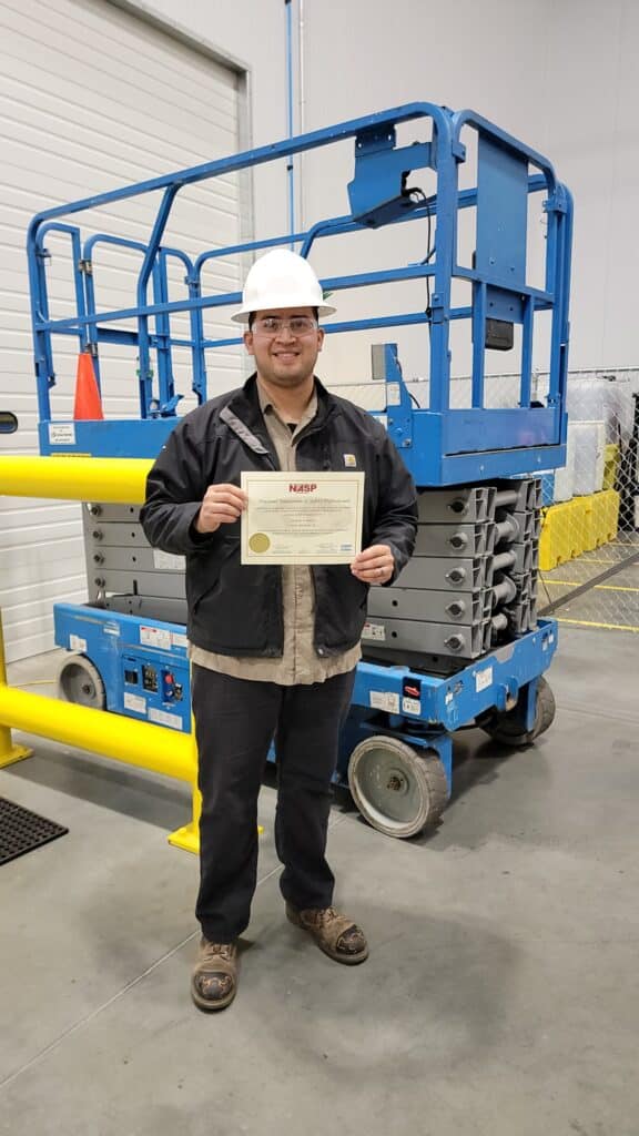 A man in a white hard hat stands in front of a blue scissor lift and holds a training certificate for upskilling employees with both hands.