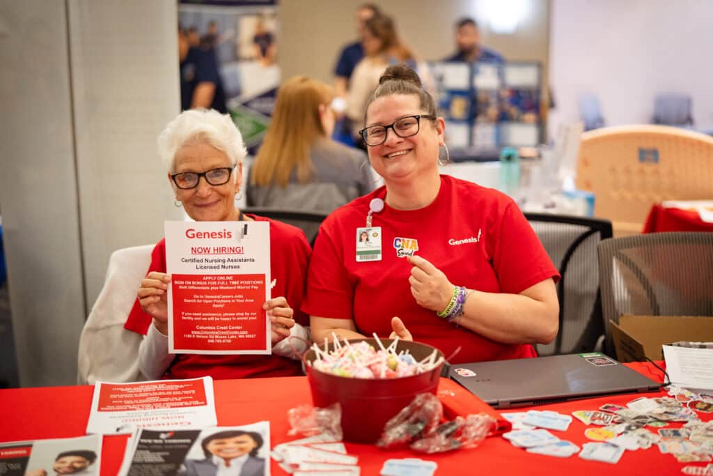 Two ladies at the BBCC Job Fair. Lady on the left with white short hair holding a &quot;Now Hiring&quot; flyer. To her right is a lady with brown hair in a bun holding a &quot;CNA squad&quot; sticker.