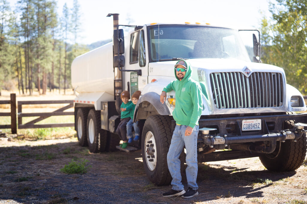A man with a green hoodie smiling at the camera while leaning against a white utility truck. Two kids in the background sitting on the side of the truck