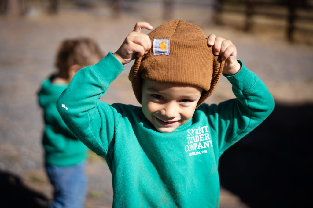 A child with a green hoodie smiling at the camera with playing with his brown hat