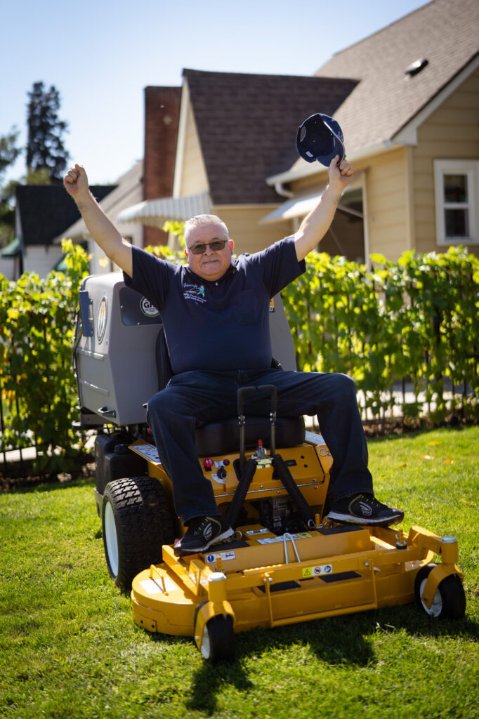 A gentleman with a navy blue hat and t-shirt sitting on a lawnmower with both hands up in the air, one hand holding up his hat in excitement
