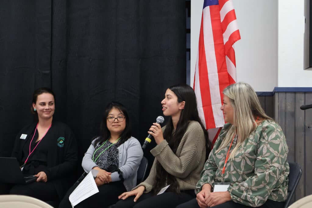 A group of 4 women in front of a stage doing a presentation. A woman wearing a olive green sweater is holding a mic