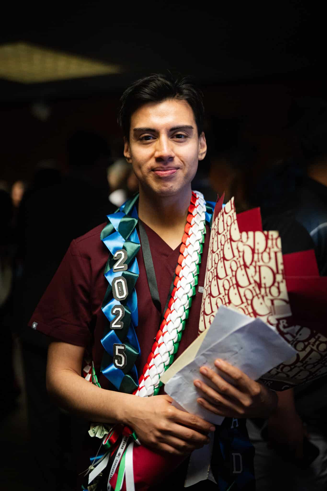A male nurse in red scrubs wears a 2025 sash and carries a bouquet of flowers.