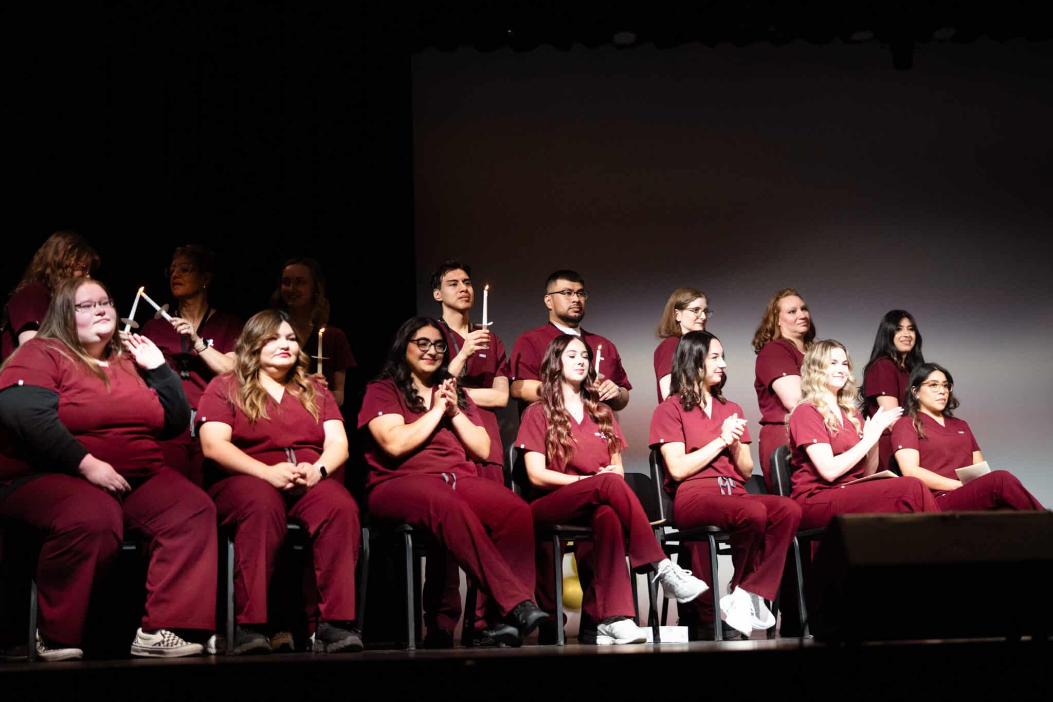 Nursing graduates in maroon scrubs clap on stage during their pinning ceremony.