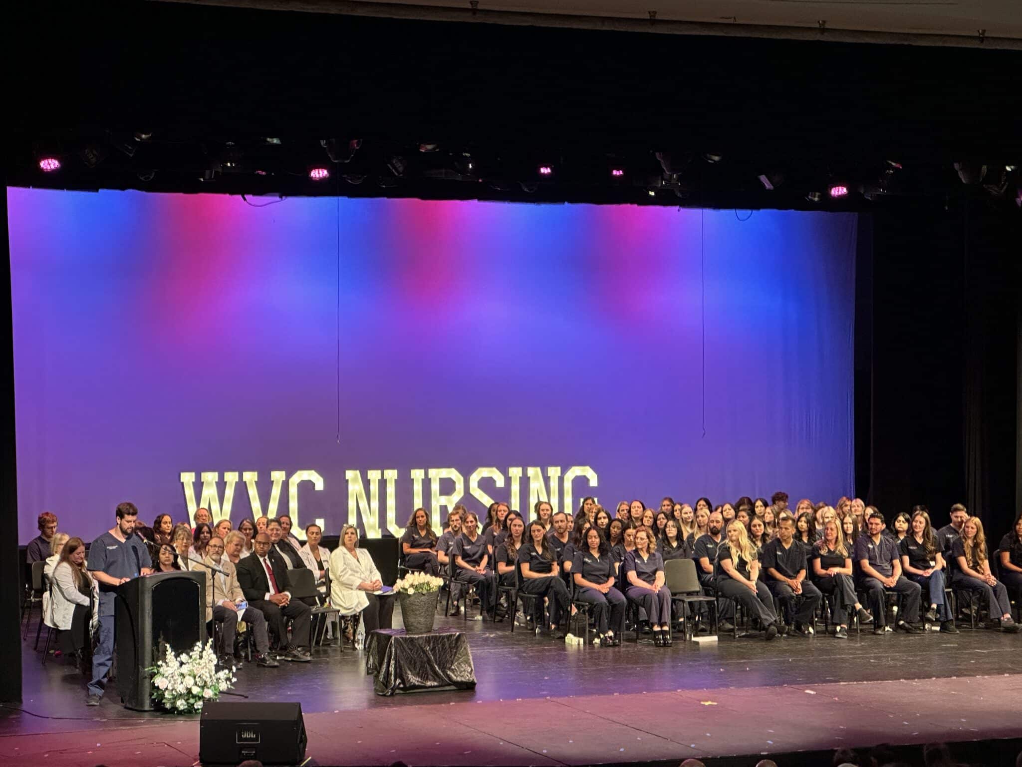 A stage filled with graduating nurses seated in folding chairs with big white letters behind them that say WVC Nursing against a blue and purple backdrop.