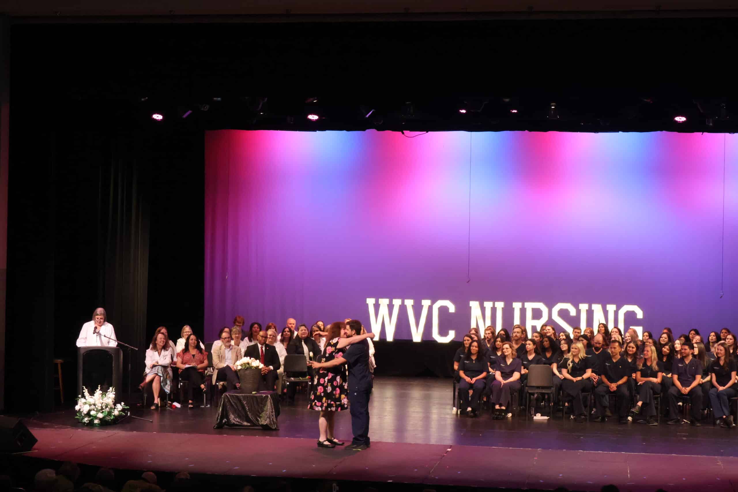 A male RN student hugging female family member in front of a stage filled with other classmates and staff