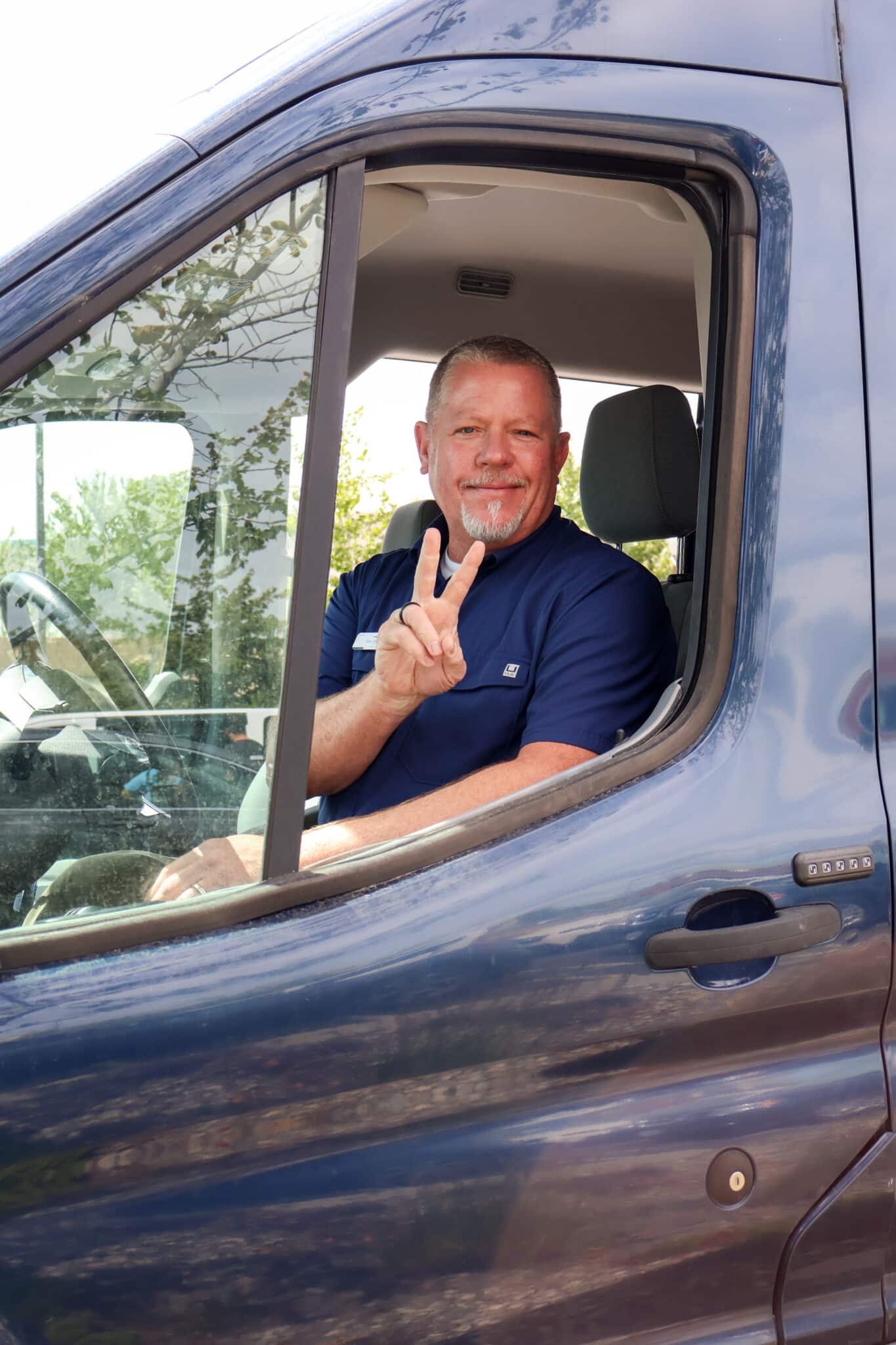 A smiling man flashes a peace sign through the window of a van.