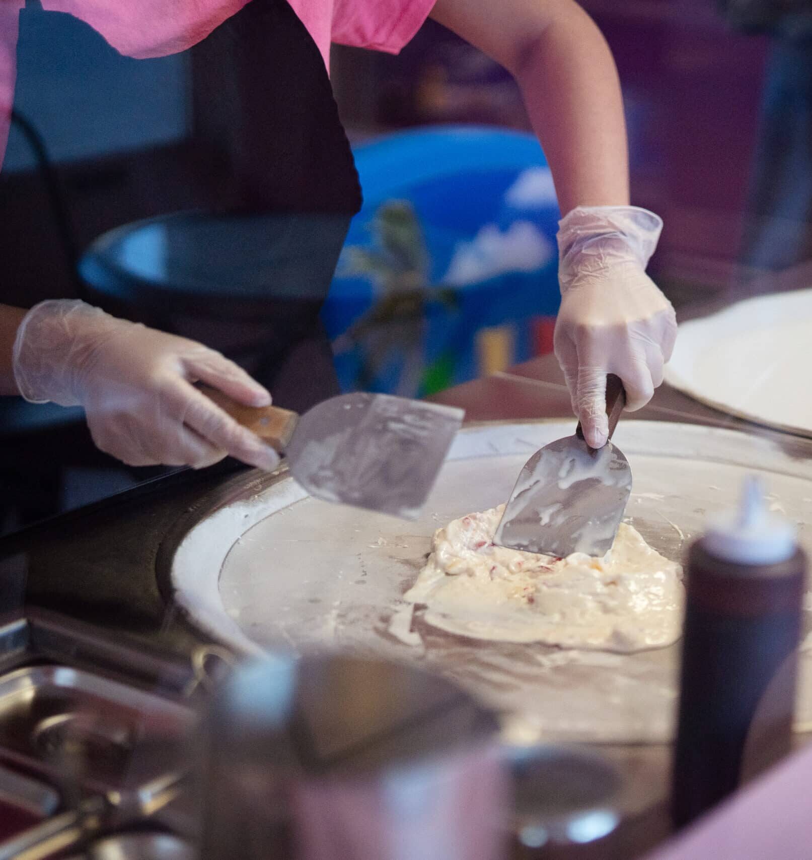 A food service worker wearing plastic gloves uses two metal spatulas to spread ice cream across a circular surface.